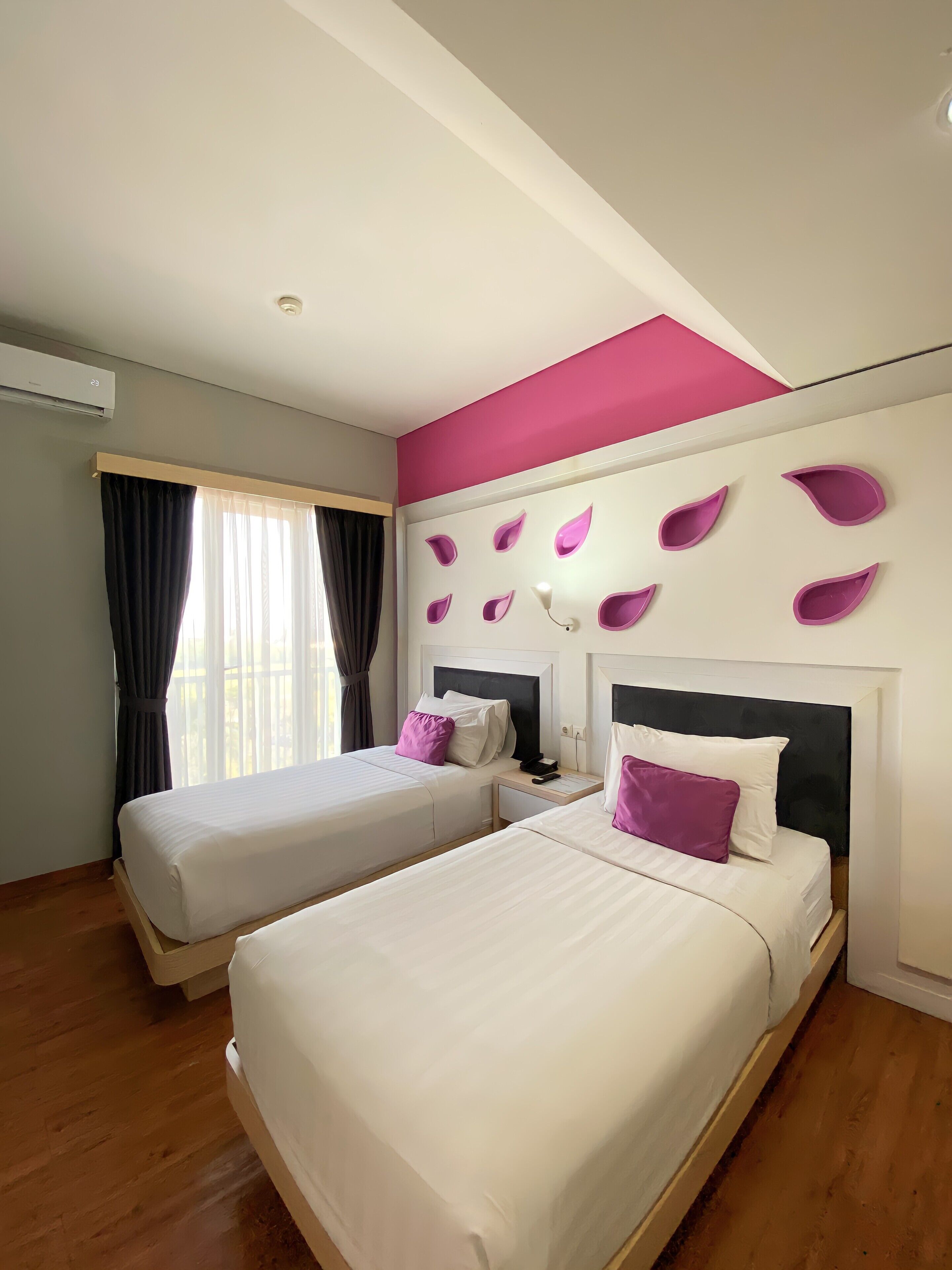A hotel room with two beds, a large window with curtains, a ceiling air conditioner, and a wall with decorative purple shapes.