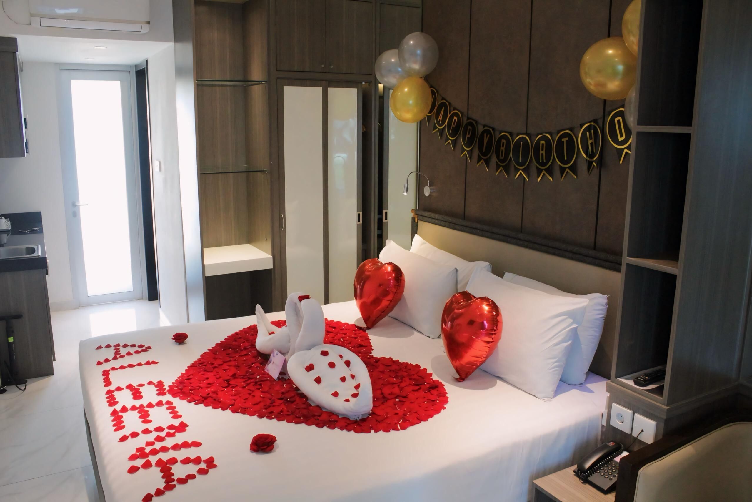 Quarto de hotel com uma cama decorada com pétalas de rosa, uma decoração de cisne e almofadas em formato de coração.