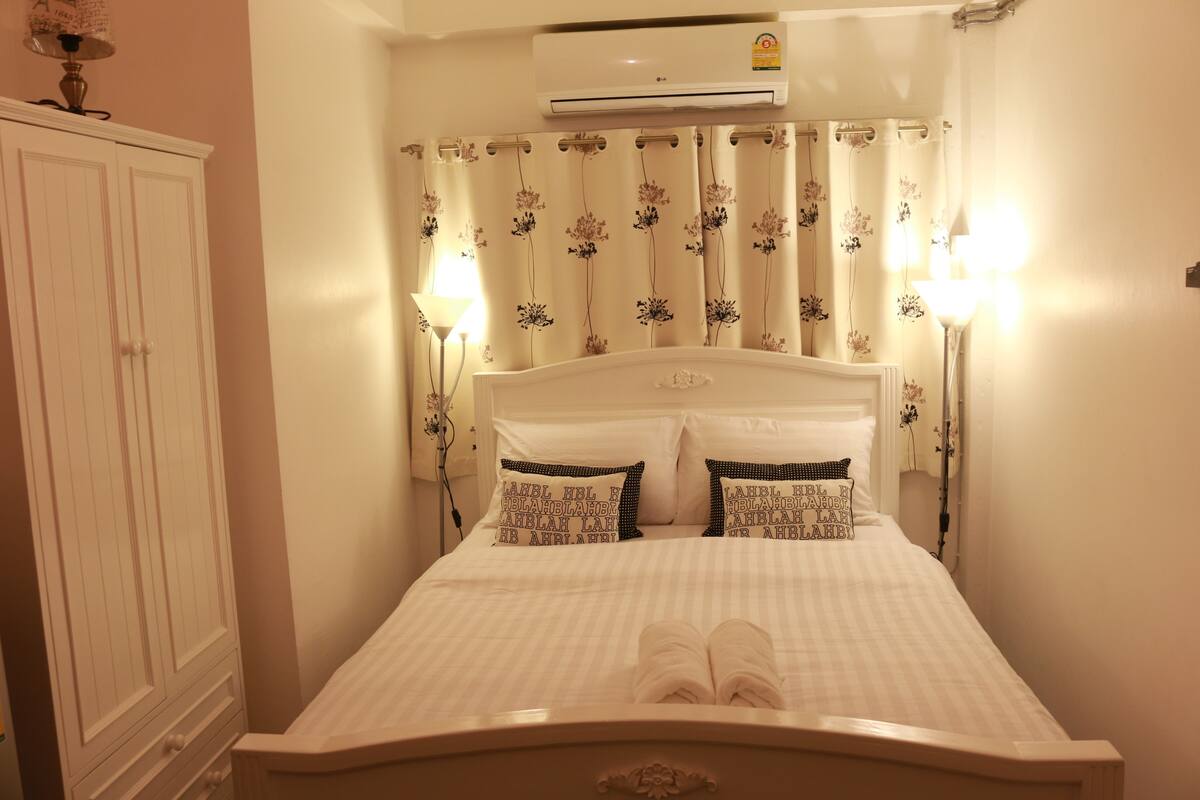 A neatly made bed with pillows and a folded towel, a white wardrobe, and a wall-mounted air conditioner.
