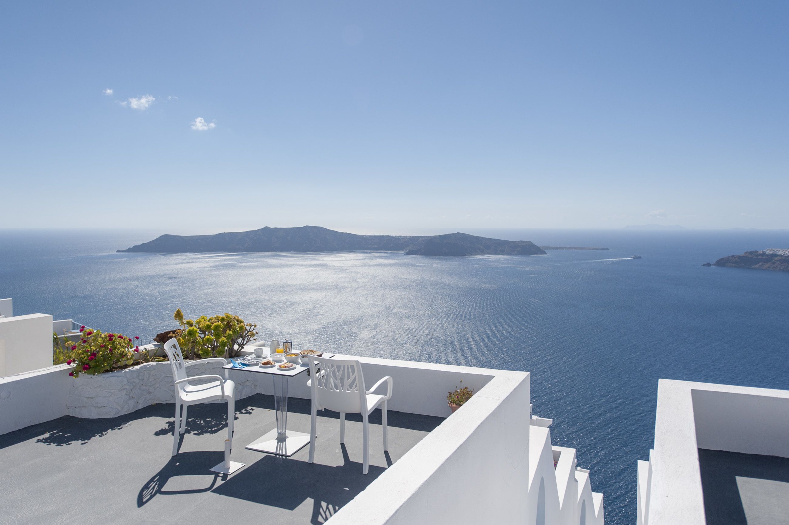 A terrace with white furniture overlooking a clear blue sea and distant islands.