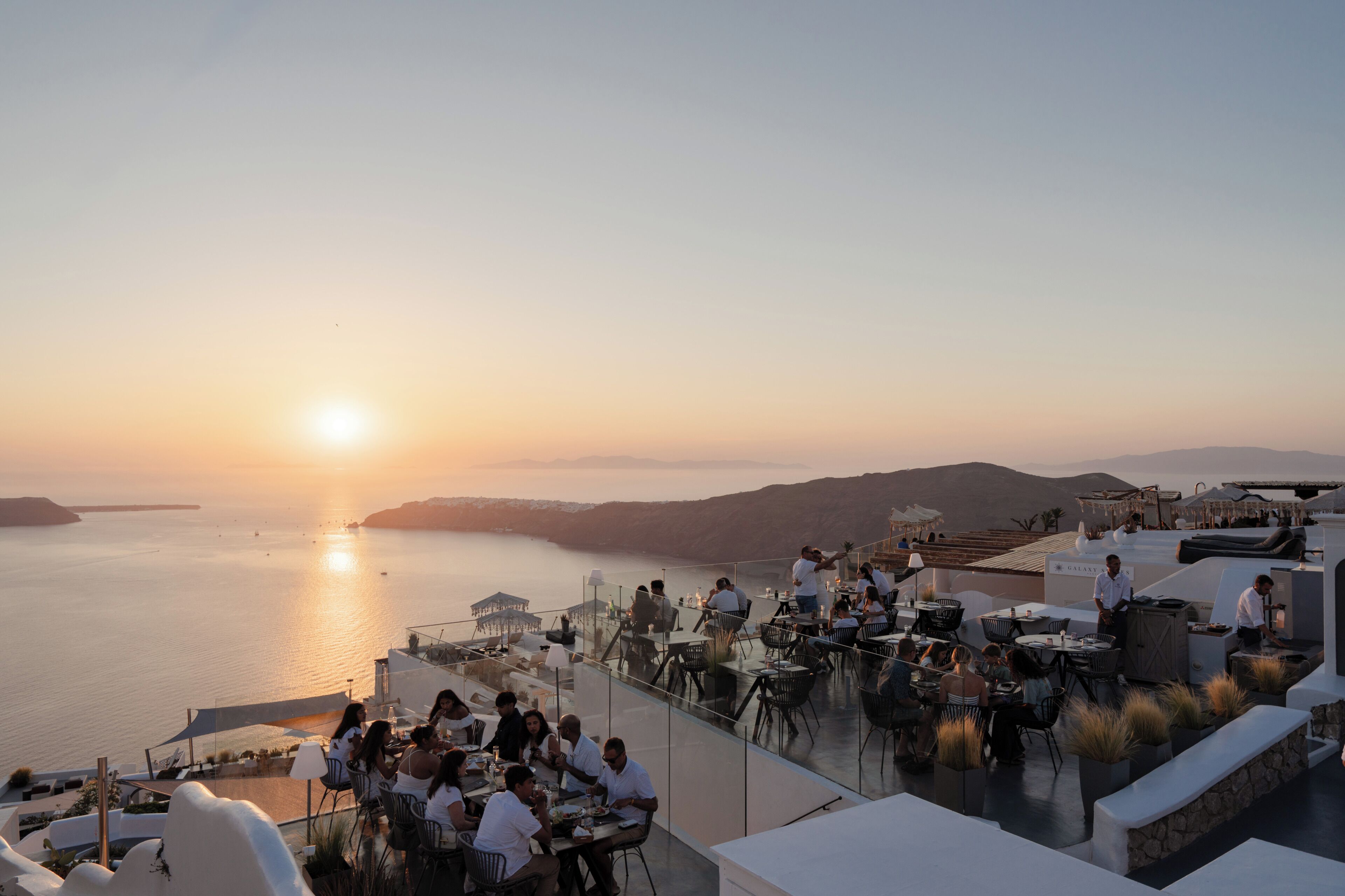 A rooftop dining area with tables and chairs, overlooking a body of water and distant hills during sunset.