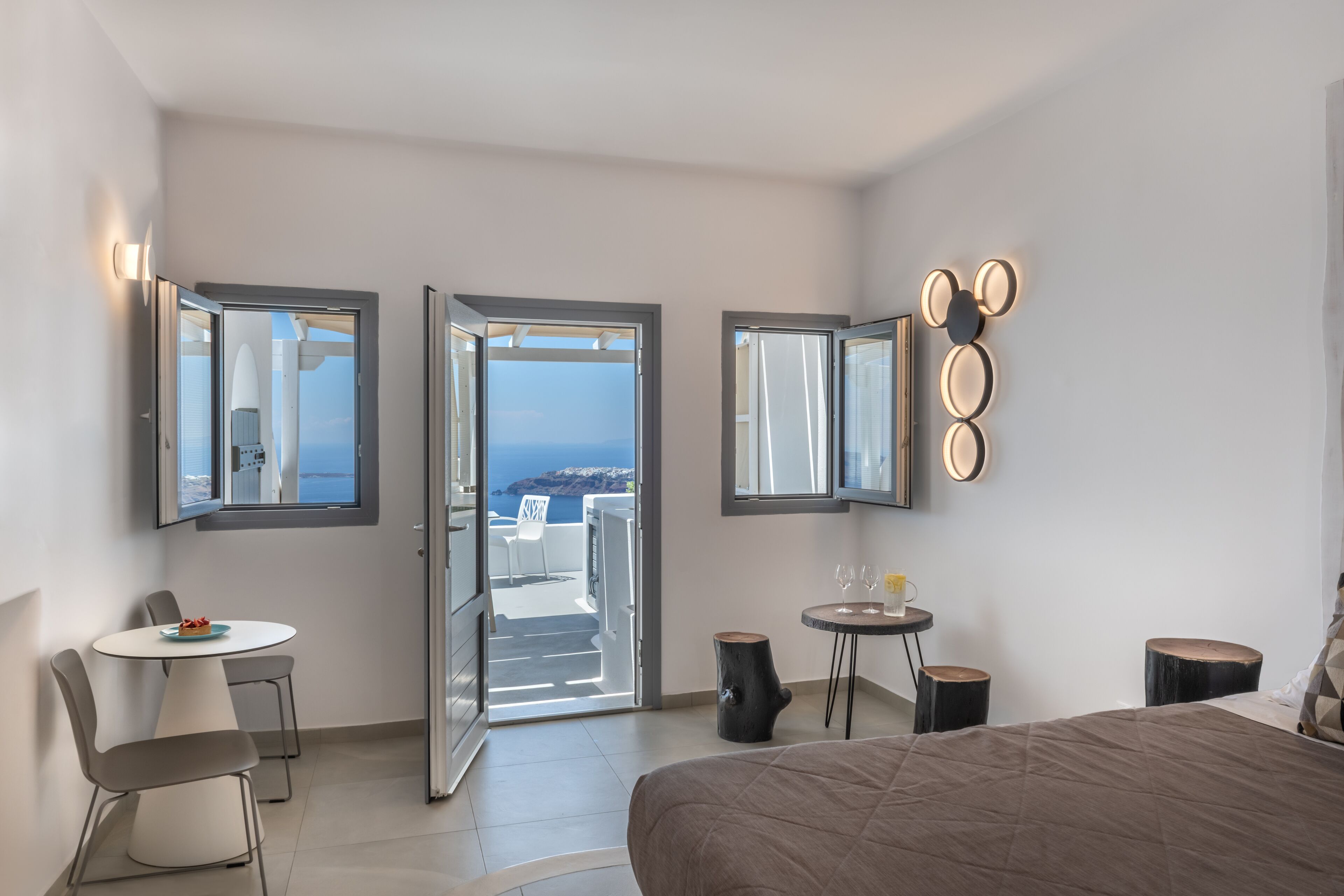 A modern hotel room with a bed, a dining table with chairs, a small round table with a drink, and a balcony with a view of the sea.