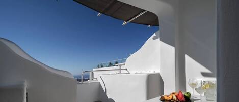 A modern outdoor balcony with a clear view of the sea, a table with fruits and drinks, and a white architectural design.
