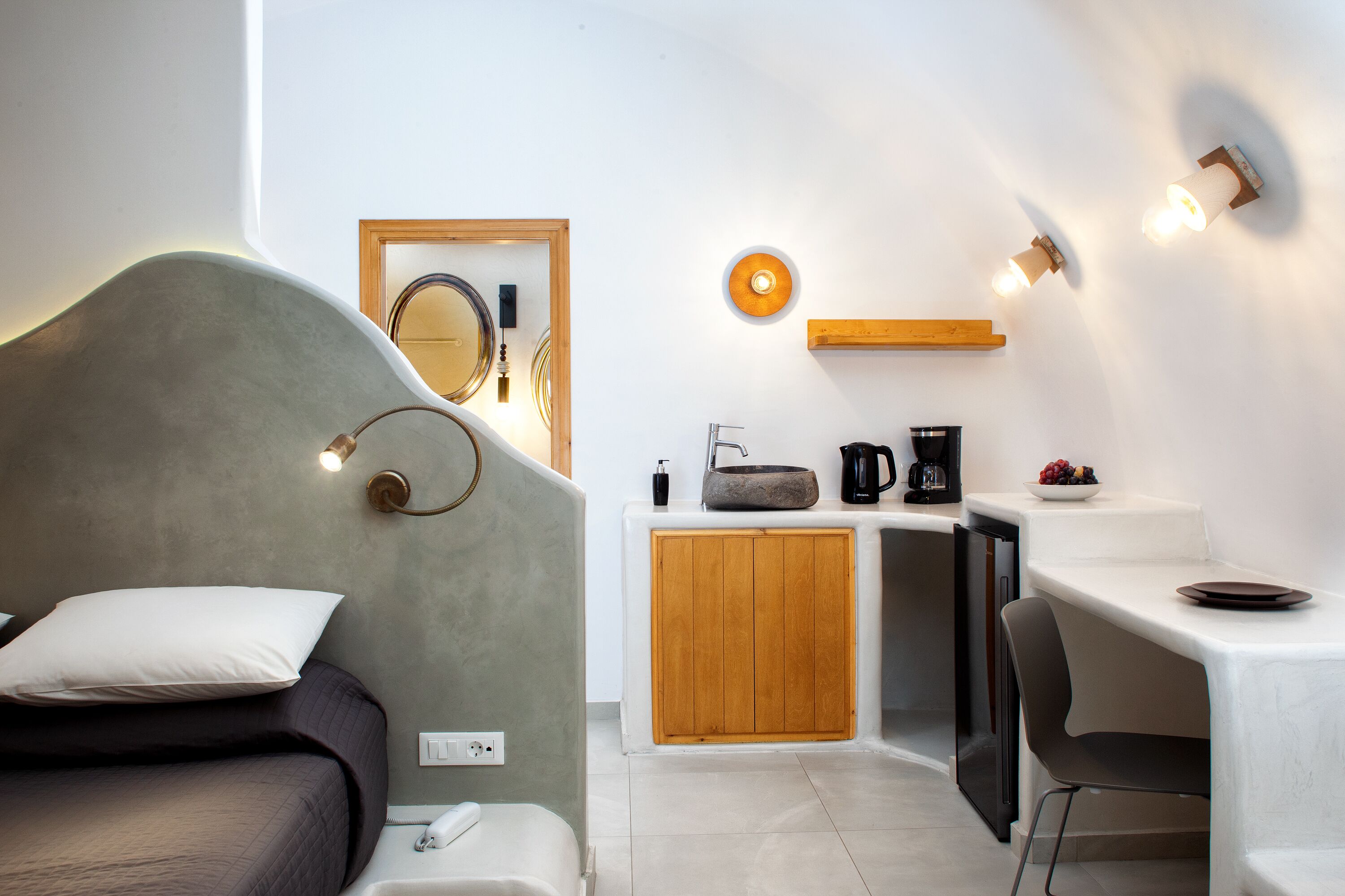 A modern bedroom with a bed, bedside table, and a small desk area with a coffee maker and fruit bowl.