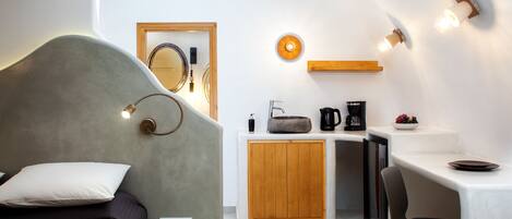 A modern bedroom with a bed, bedside table, and a small desk area with a coffee maker and fruit bowl.