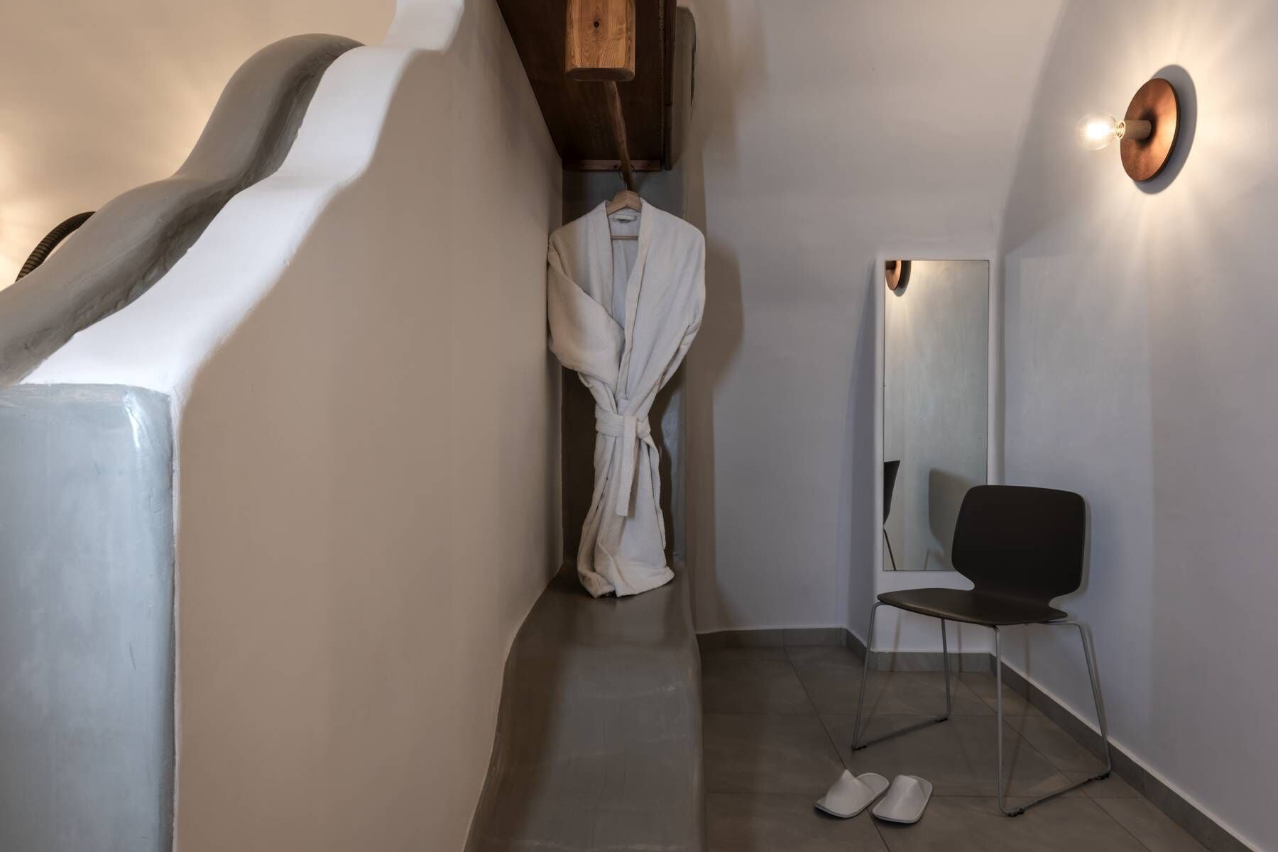 A narrow hallway with a white robe hanging on a rack, a black chair, and a mirror on the wall.