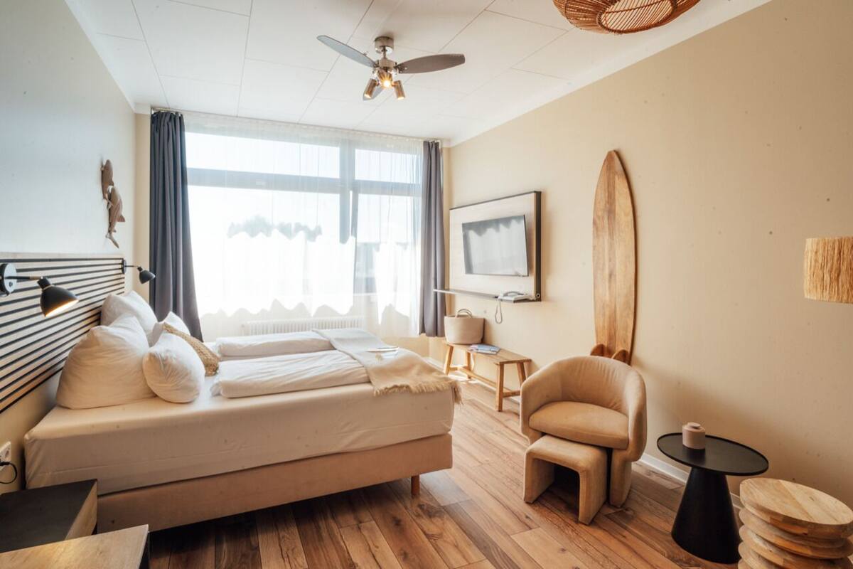 Ein modernes Hotelzimmer mit Bett, Sessel, kleinem Tisch, Surfbrett und Deckenventilator.