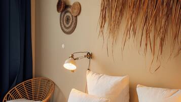 A neatly made bed with white linens, a woven wall hanging, a wall-mounted lamp, and a wicker chair.
