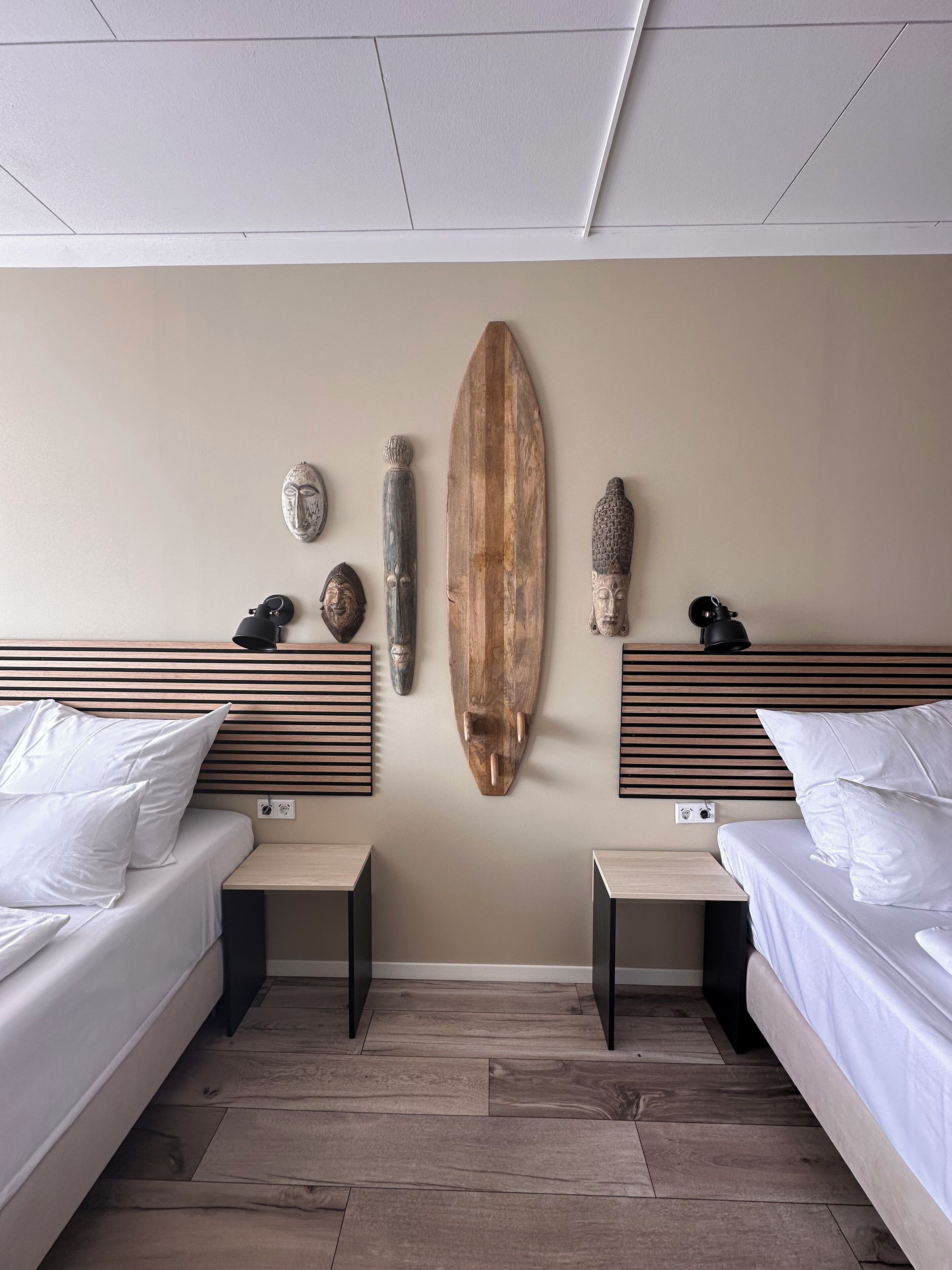 Une chambre d’hôtel moderne avec deux lits, des têtes de lit en bois et une planche de surf accrochée au mur.