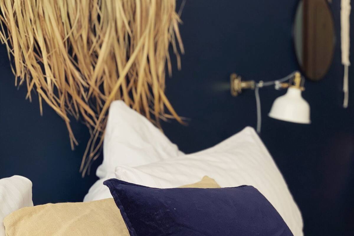 Ein Bett mit weißer Bettwäsche und zwei Kissen, eines beige, eines dunkelblau, in einem Raum mit dunklen Wänden und einer Pendelleuchte.