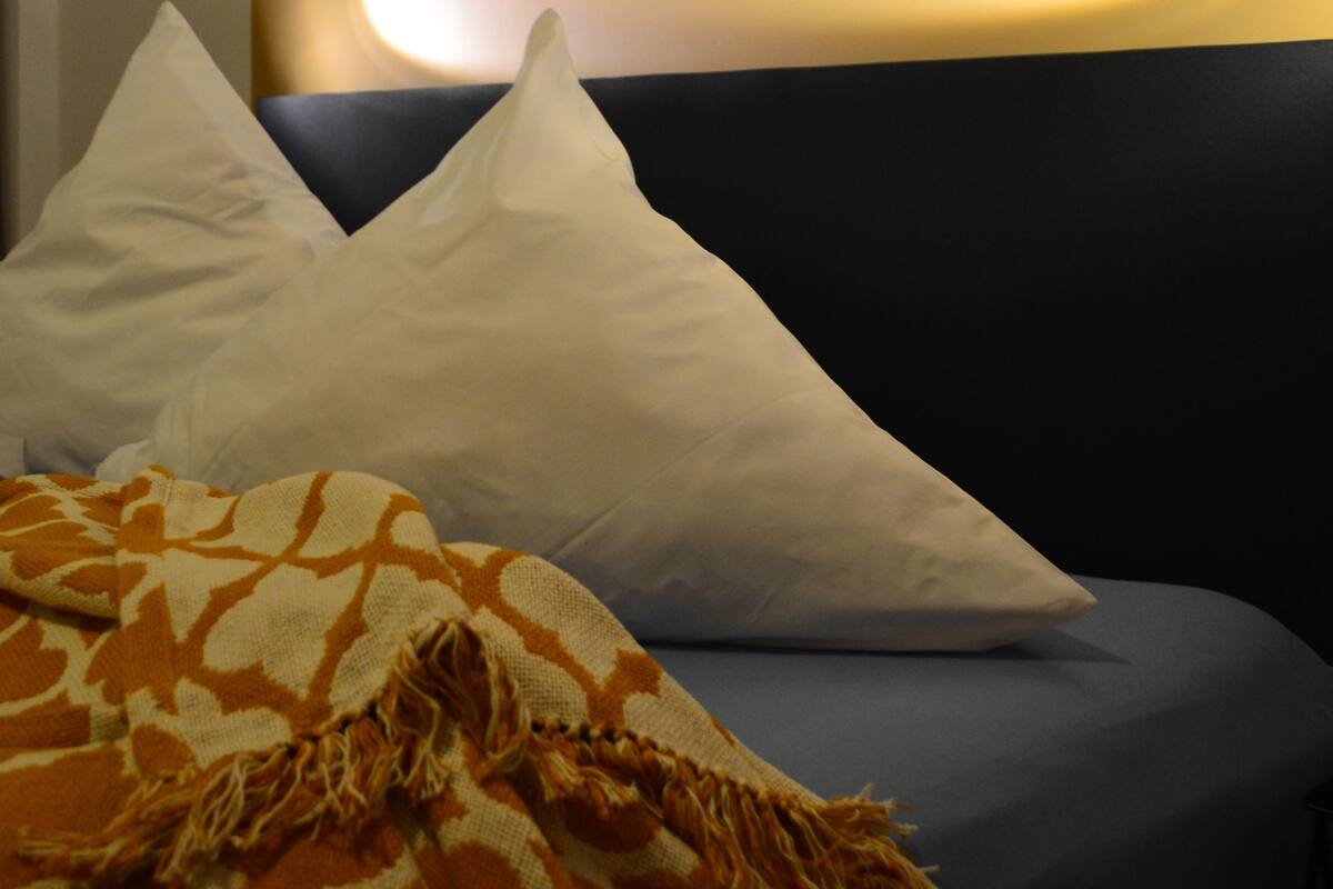 Ein Bett mit weißen und orangefarbenen Kissen, einem dunklen Kopfteil und einem Gemälde von stürmischer See an der Wand.