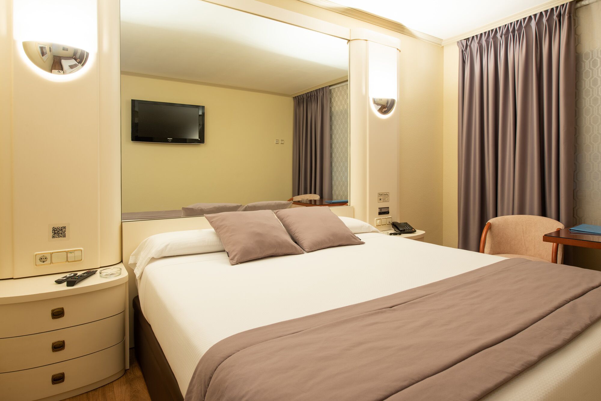 A hotel room with a large bed, bedside tables, a desk, a chair, a TV, and curtains.