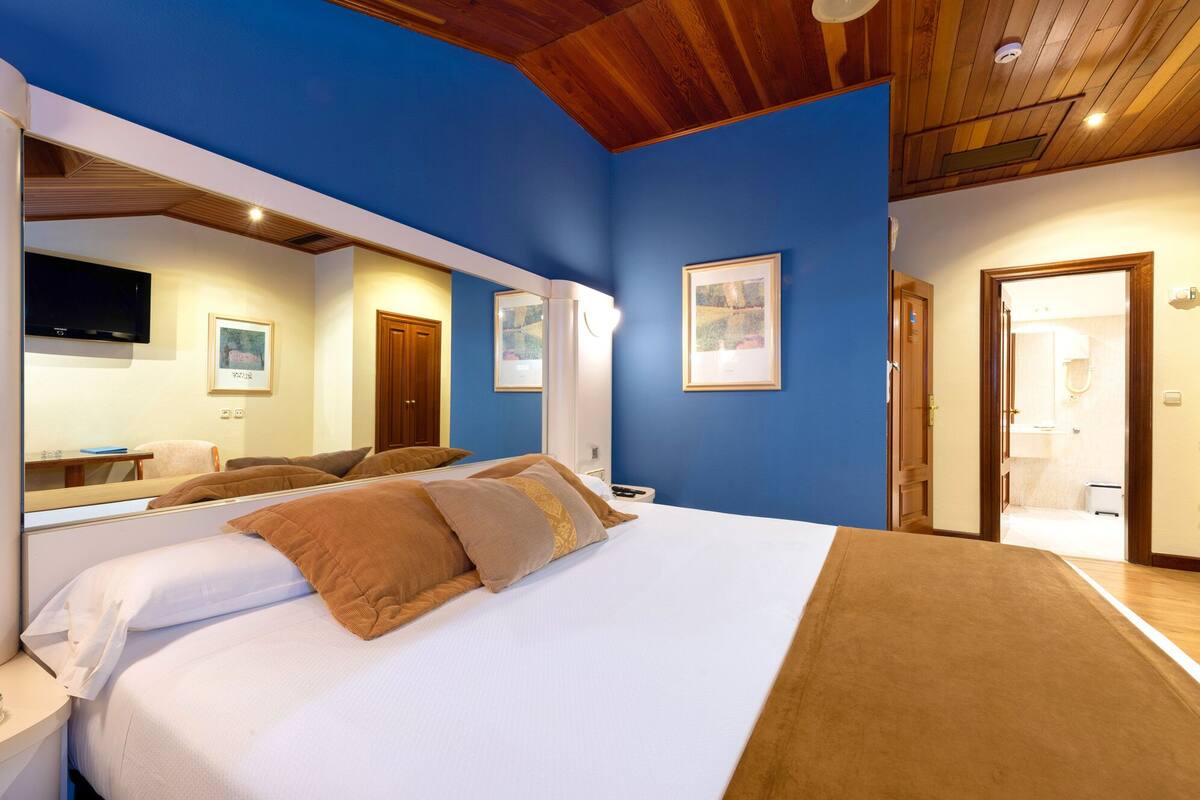 A bedroom with a large bed, a wooden ceiling, a blue wall, and a door leading to a bathroom.