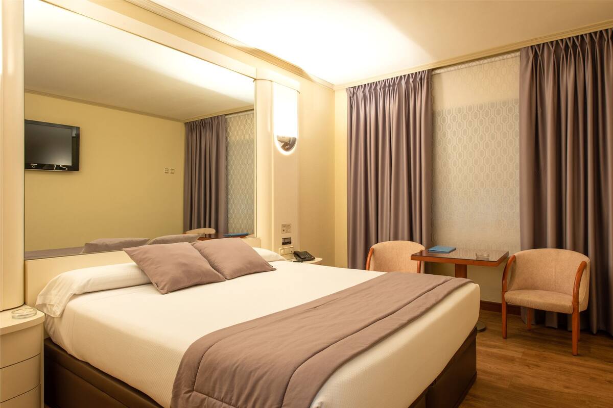 A hotel room with a large bed, a desk, a chair, a TV, and curtains.