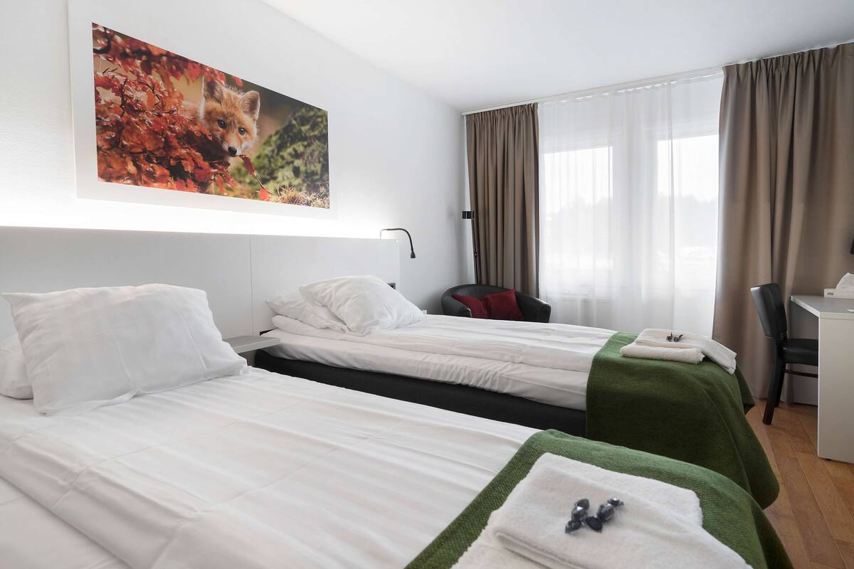 A hotel room with two beds, a desk, a chair, and a large window with curtains.