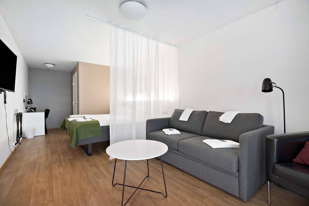 A modern living room with a grey sofa, a small white table, a black floor lamp, and a green blanket on a bed.