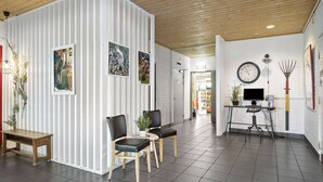 Ein modernes Interieur mit Holzboden, weißen Wänden und Fliesenboden. Zwei Stühle und ein kleiner Tisch mit einer Pflanze sind vorhanden. Auf einem Schreibtisch befinden sich eine Uhr und ein Computermonitor.