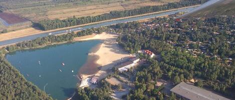 An aerial view of a lakeside area with a small beach, surrounded by dense forests and residential buildings.