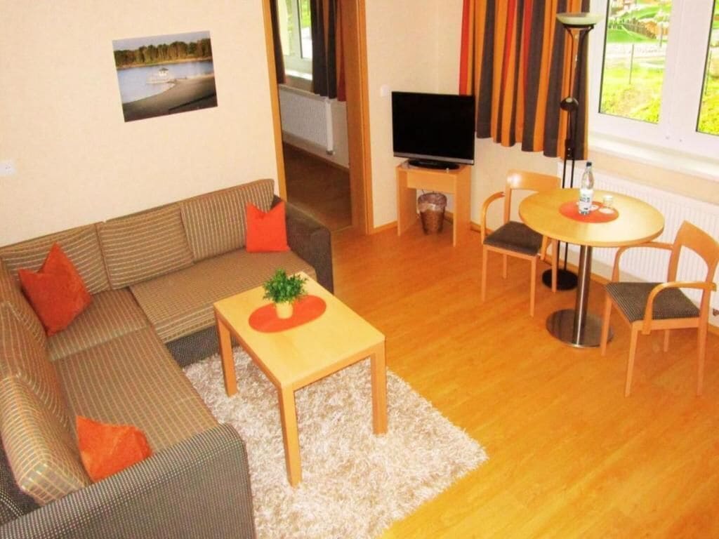 A living room with a sofa, a small table with a plant, a TV on a stand, and a dining area with chairs and a table.