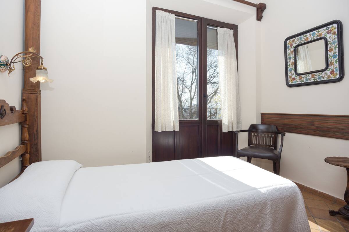 Un dormitorio con una cama, una silla, una mesita, una ventana con cortinas y un espejo en la pared.