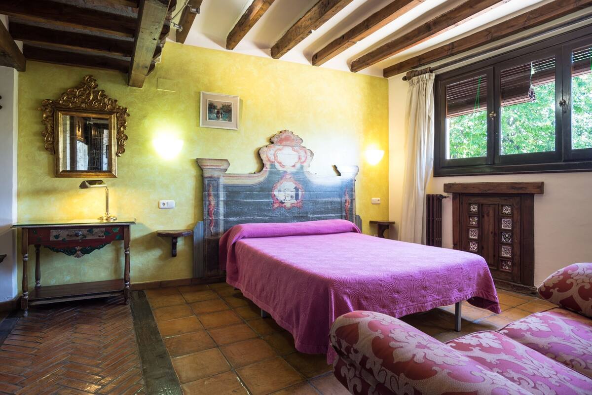 Un dormitorio con cama, mesita de noche, espejo y ventana con cortinas.