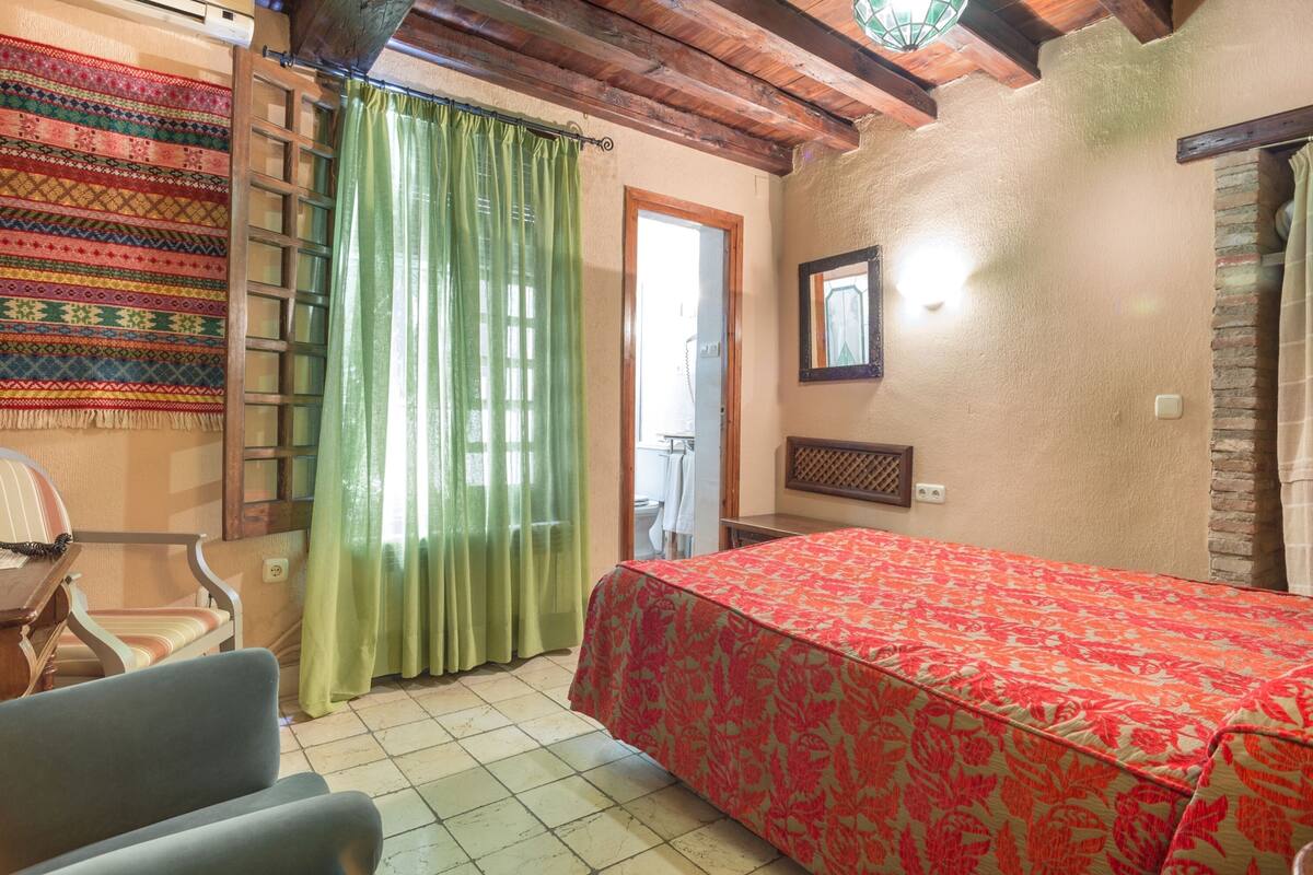 Una habitación con una cama, una silla, una mesa de madera, una cortina verde y un tapiz de pared estampado.
