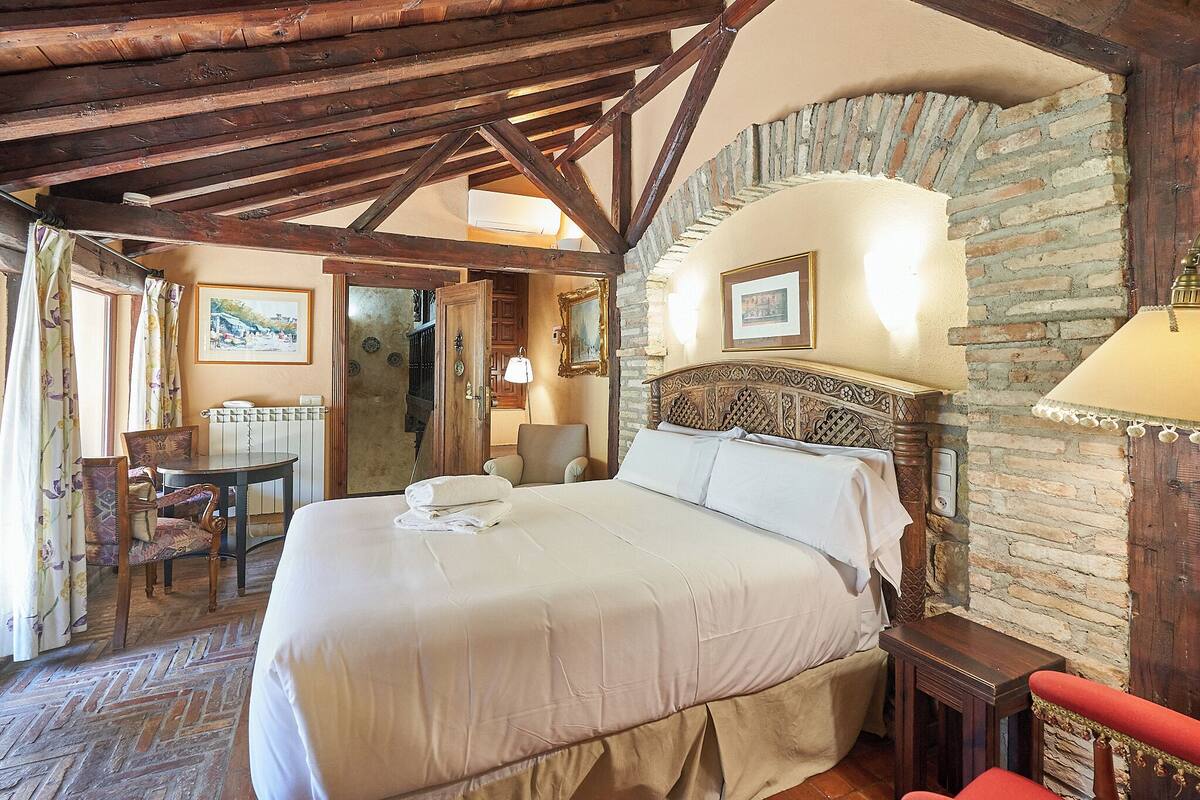 A hotel room with a large bed, wooden ceiling beams, stone walls, a small table, and a chair.