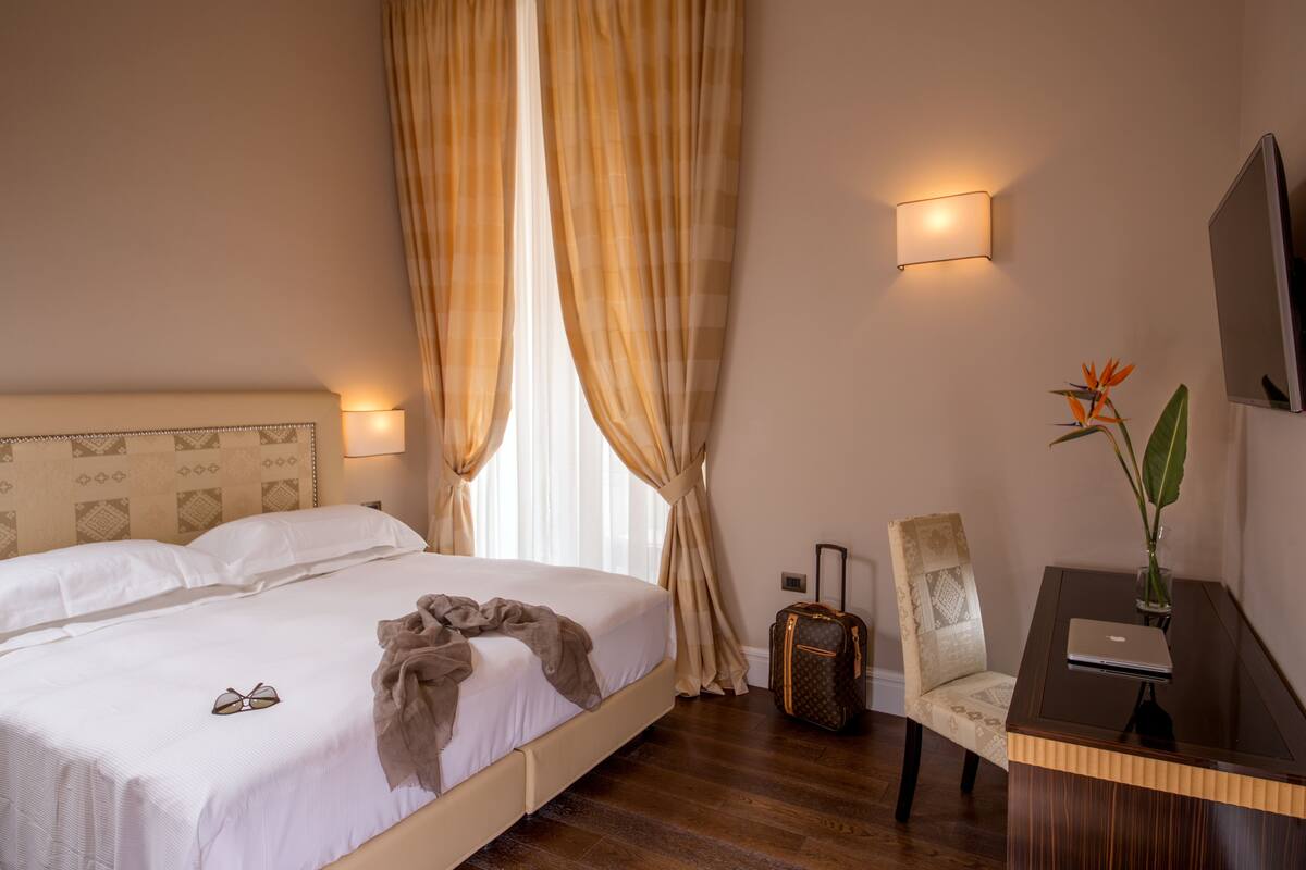 A hotel room with a bed, a desk, a chair, a suitcase, and a plant.