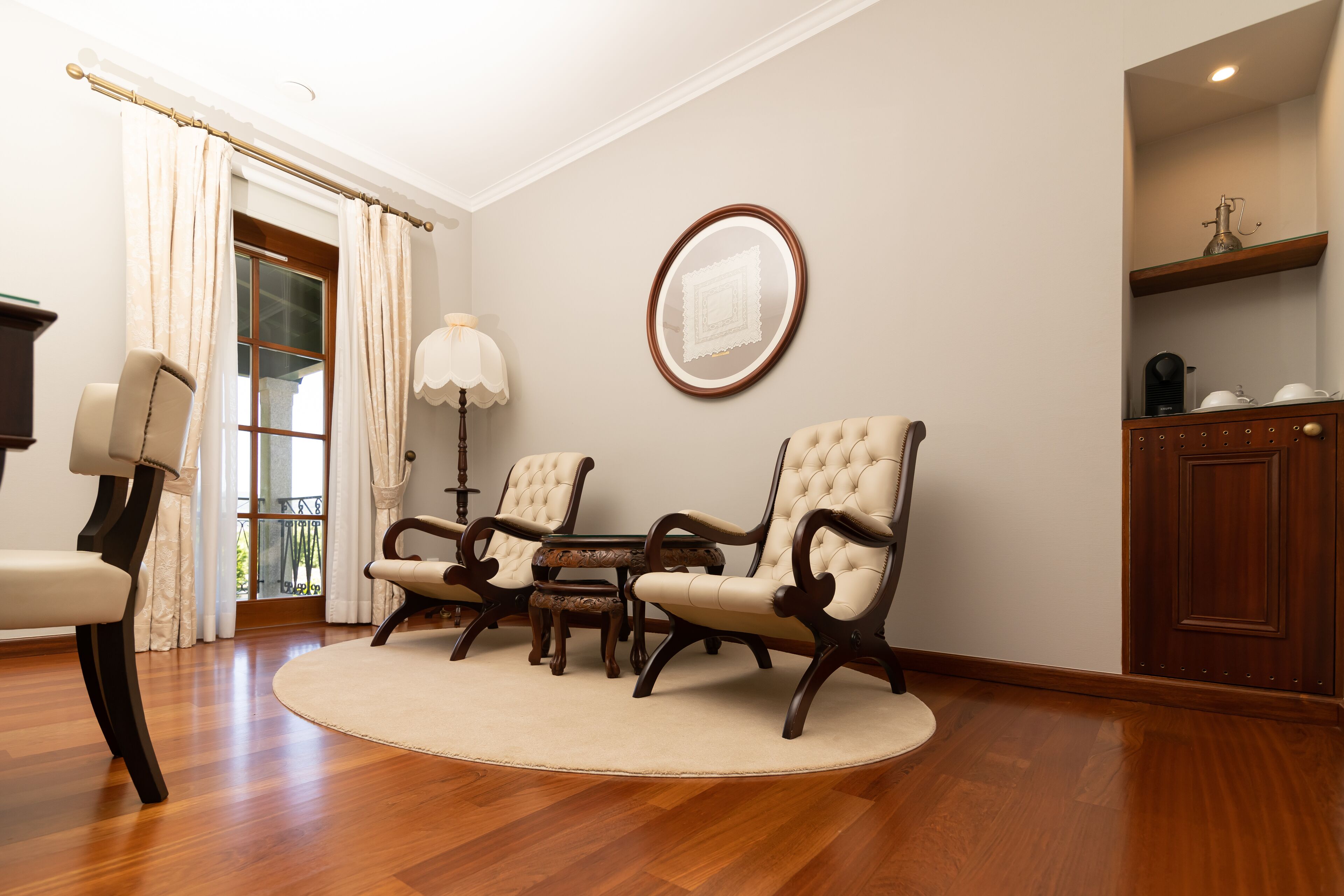 A room with wooden flooring, a round rug, two upholstered chairs, a small table, a mirror on the wall, and a door with a curtain.
