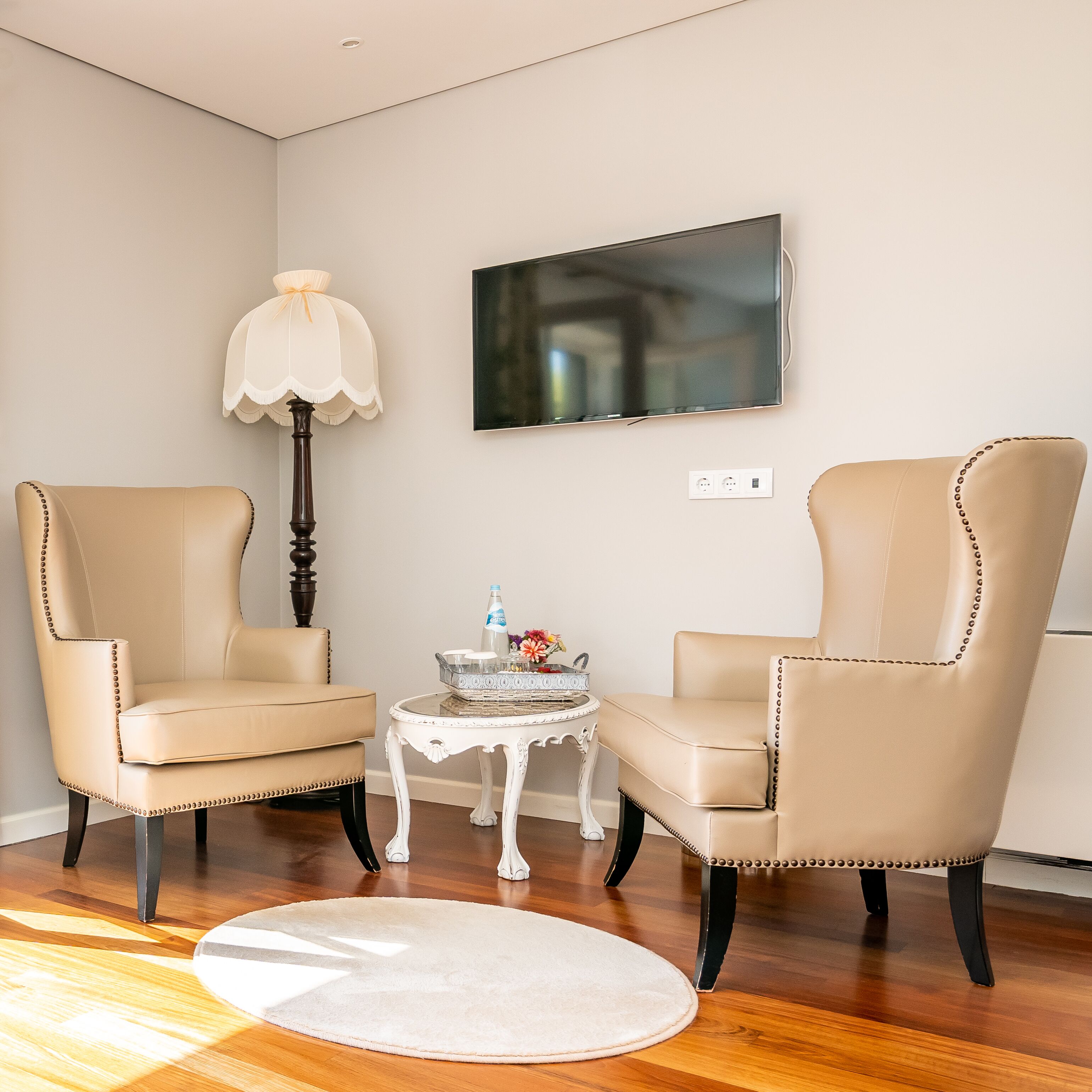 Una habitación con dos sillones beige, una mesa blanca con una lámpara y un televisor de pantalla plana montado en la pared.