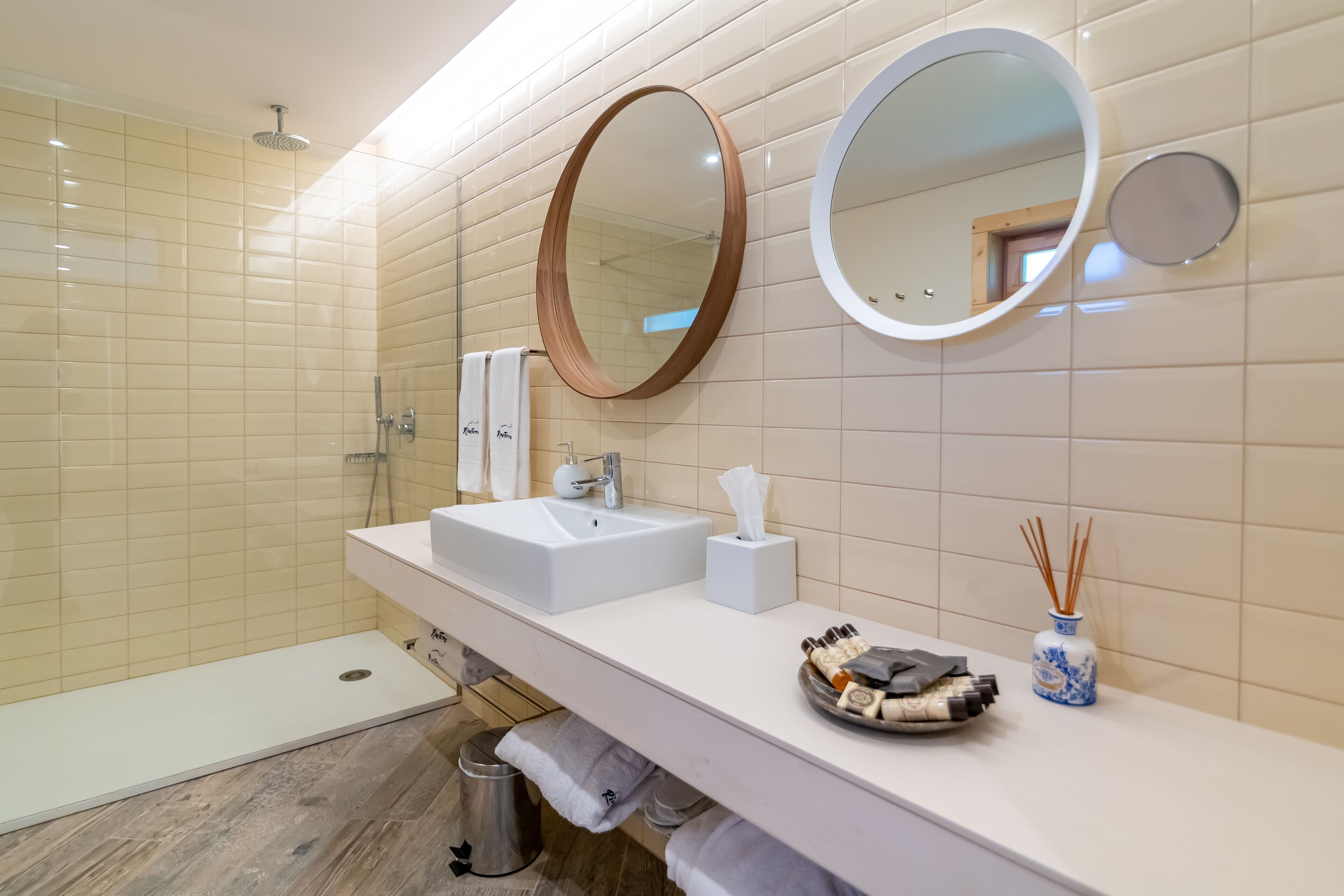 A modern bathroom with a large sink, two mirrors, a shower, and a vanity with toiletries.