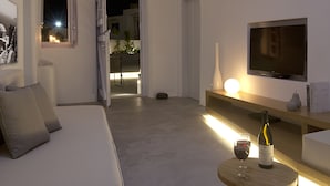 A modern living room with a sofa, a TV, a small table with a bottle and glass, and a lit window. - Anemolia Villa (Santorini)