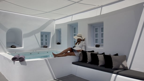 A woman sitting by a pool in a white room with windows and a bathtub. - Anemolia Villa (Santorini)