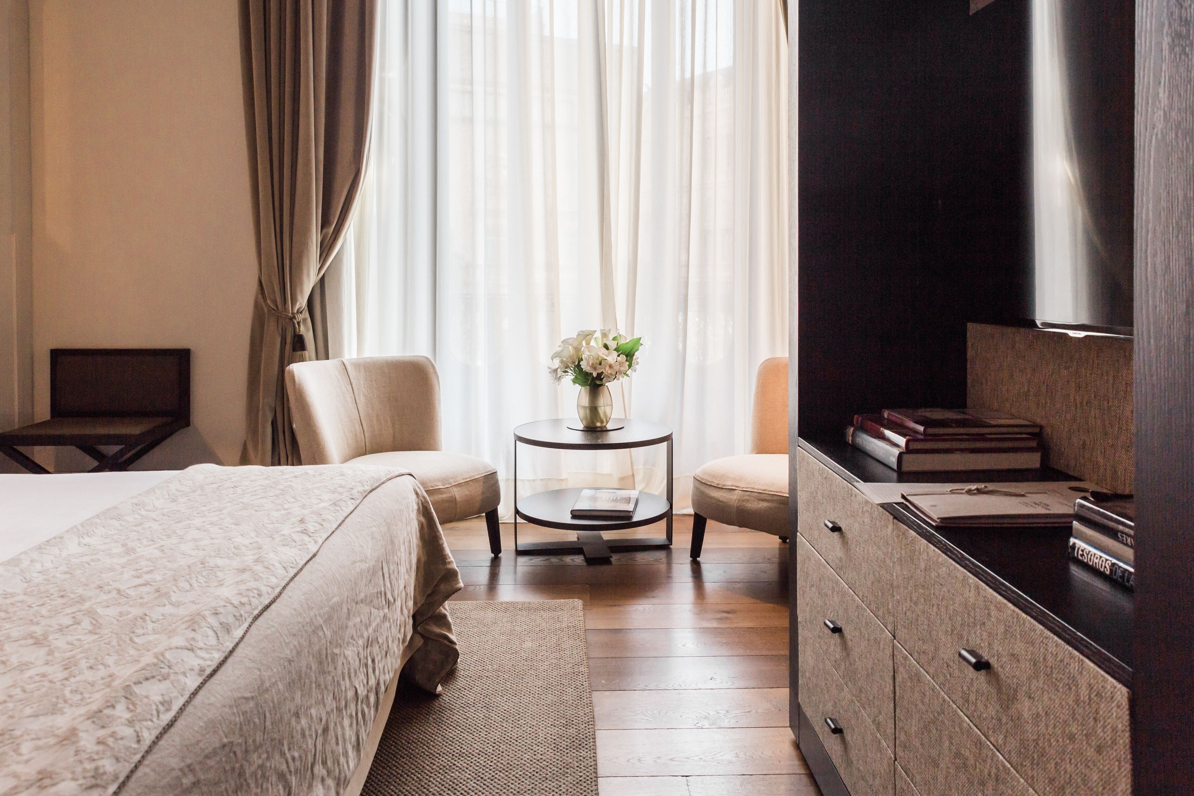 A hotel room with a bed, two chairs, a small table with a vase of flowers, a TV, and a dresser with books.