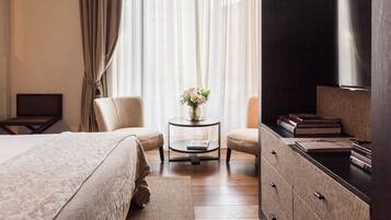 A hotel room with a bed, two chairs, a small table with a vase of flowers, a TV, and a dresser with books.