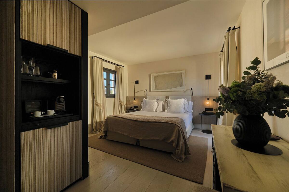 A hotel room with a bed, bedside table, lamp, and a shelf with glasses and a radio.