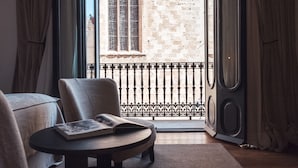A room with a view of a stone building through a window, a round table with a book, a sofa, and a speaker. - Sant Francesc Hotel Singular (Palma de Mallorca)