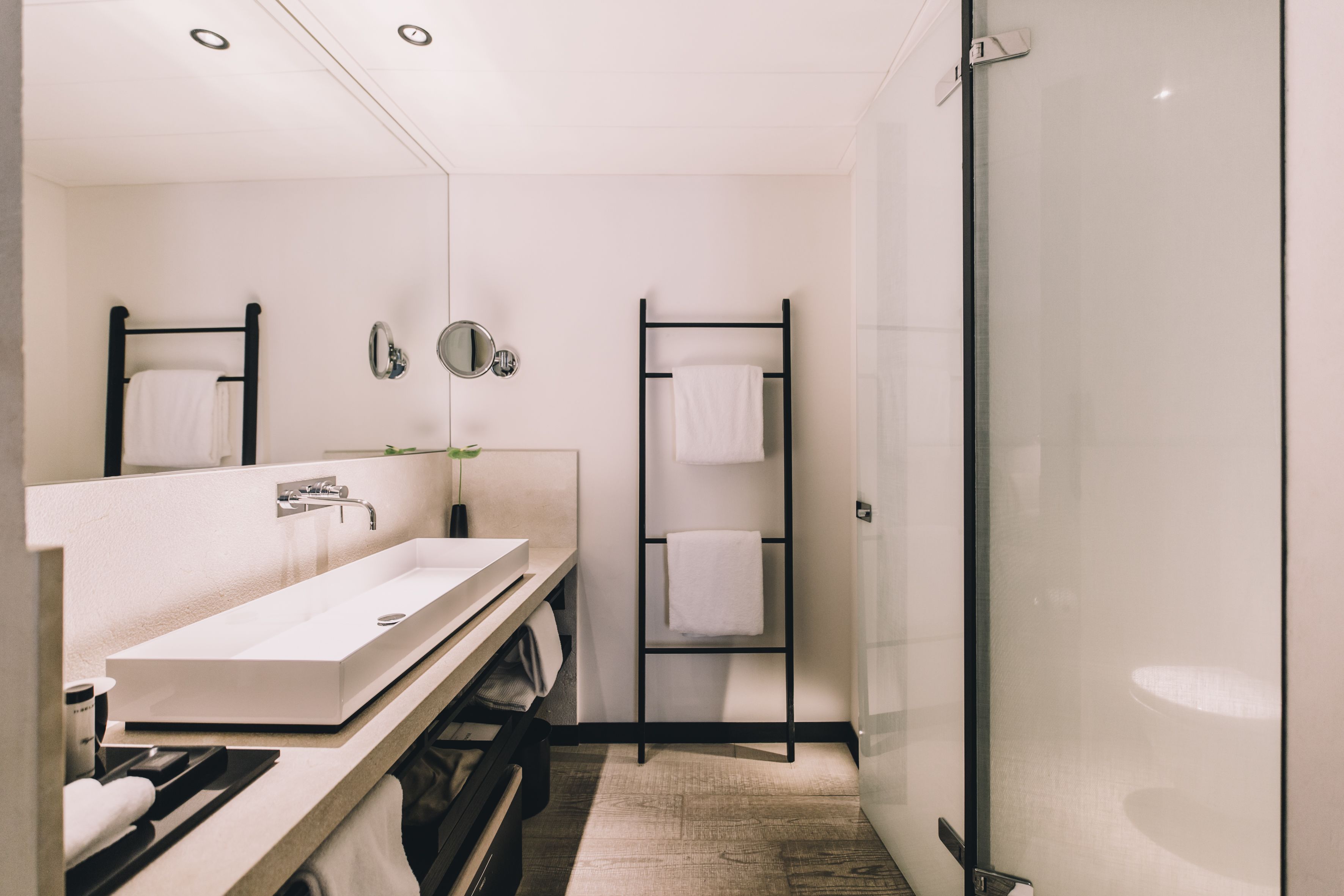 A modern bathroom with a large white sink, two mirrors, a towel rack, and a glass shower enclosure.