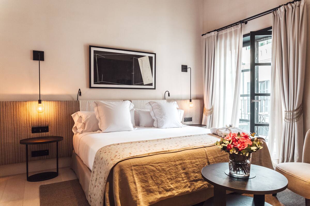 A hotel room with a bed, bedside lamps, a framed artwork, a window with curtains, and a vase with flowers on a side table.