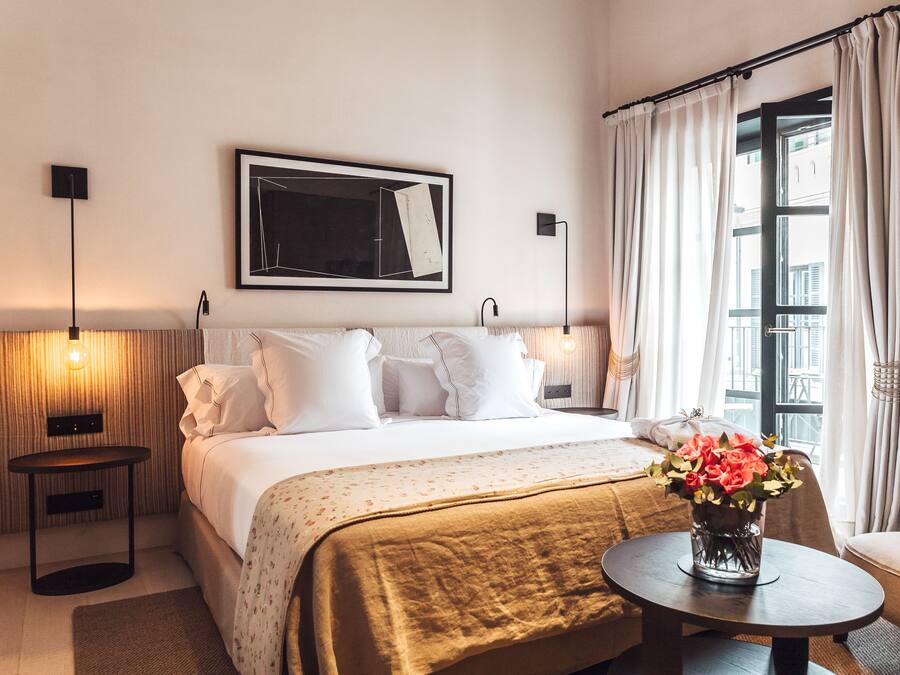 A hotel room with a bed, bedside lamps, a framed artwork, a window with curtains, and a vase with flowers on a side table.
