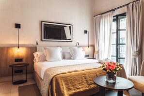 Une chambre d’hôtel avec un lit, des lampes de chevet, un tableau encadré, une fenêtre avec des rideaux et un vase avec des fleurs placé sur une table d’appoint.