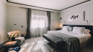 Habitación de hotel con una cama grande, una mesita de noche con lámpara, una silla, una pequeña mesa con un jarrón de flores y una ventana con cortinas.