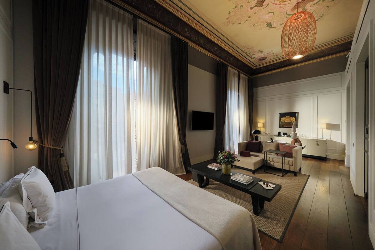 A hotel room with a large bed, a sitting area with a sofa and armchairs, a dining table, and a chandelier.