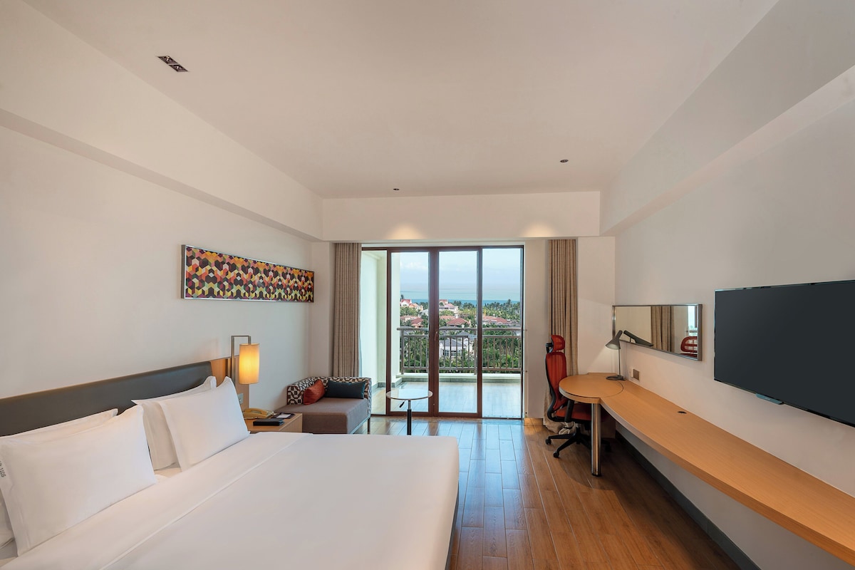 Hotelkamer met een bed, een bureau, een stoel, een televisie, een schilderij en een balkon met uitzicht.