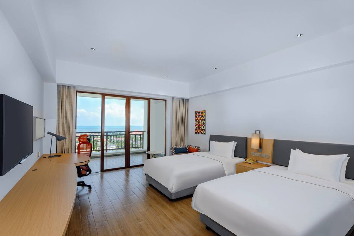 Habitación de hotel con dos camas, un escritorio, una silla, un televisor, un balcón con vistas y un cuadro en la pared.