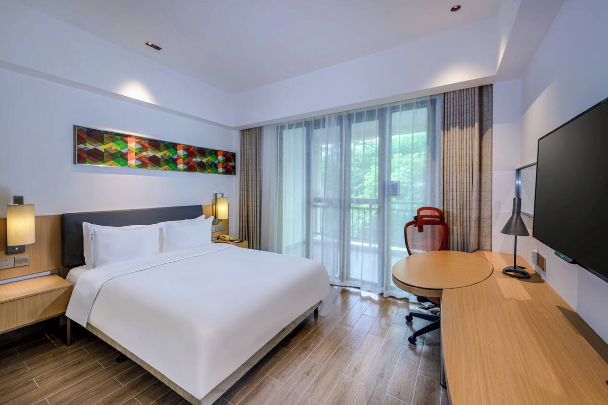 Una habitación de hotel moderna con una cama grande, un escritorio, una silla, un televisor y una ventana con cortinas.