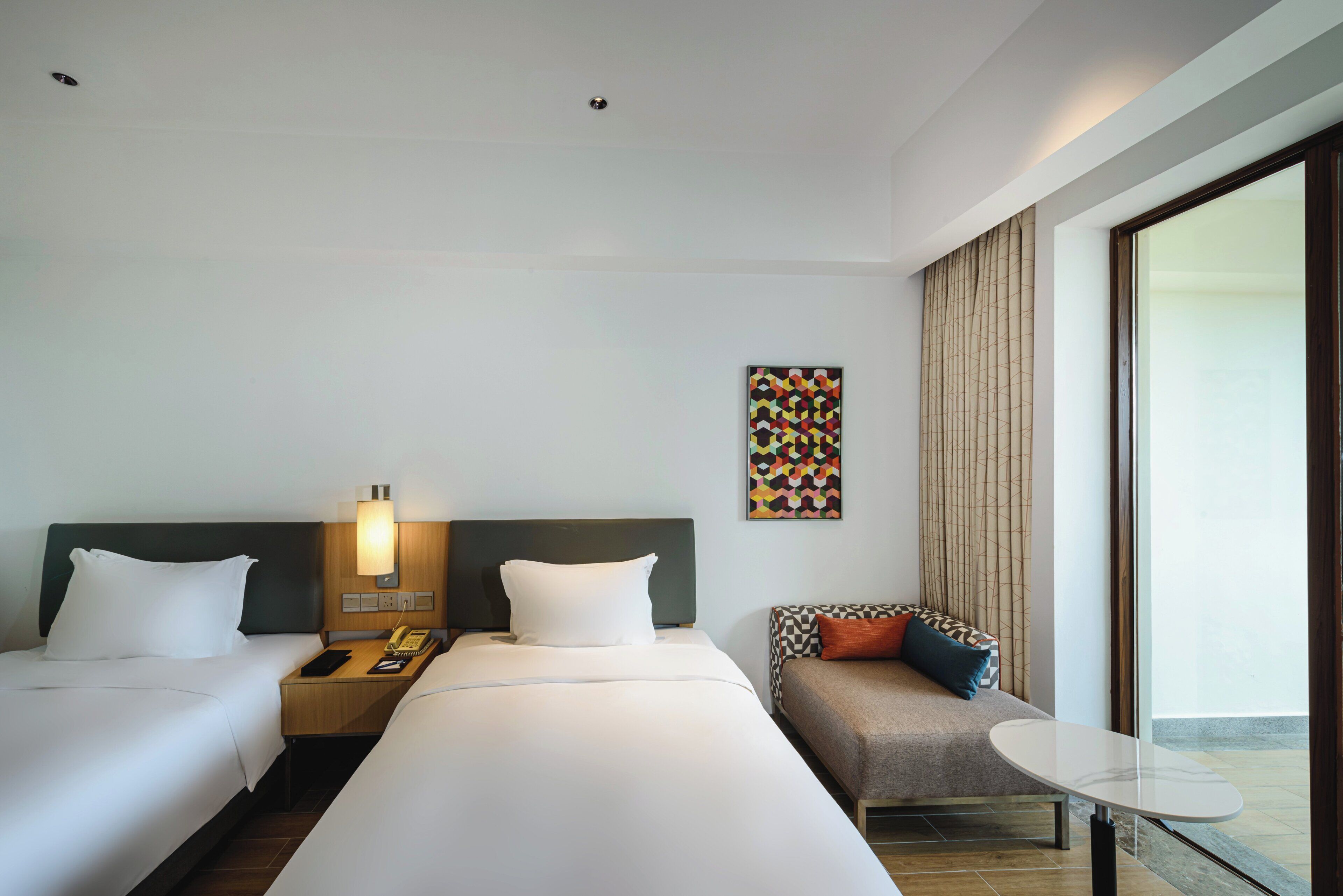 A hotel room with two beds, a desk with a lamp, a sofa, and a large window.