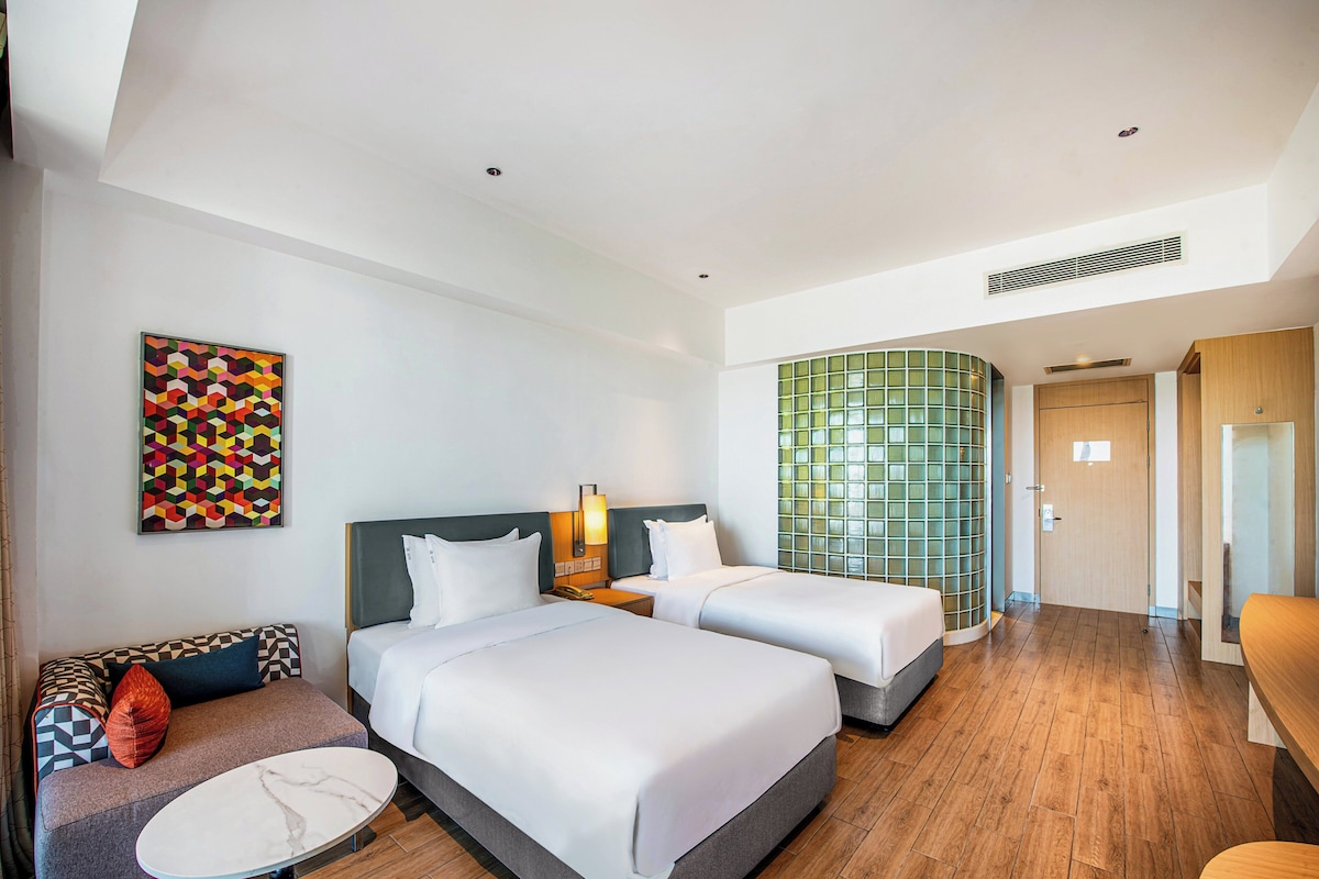 Een hotelkamer met twee bedden, een bank, een kleine tafel en een kleurrijk abstract schilderij aan de muur.