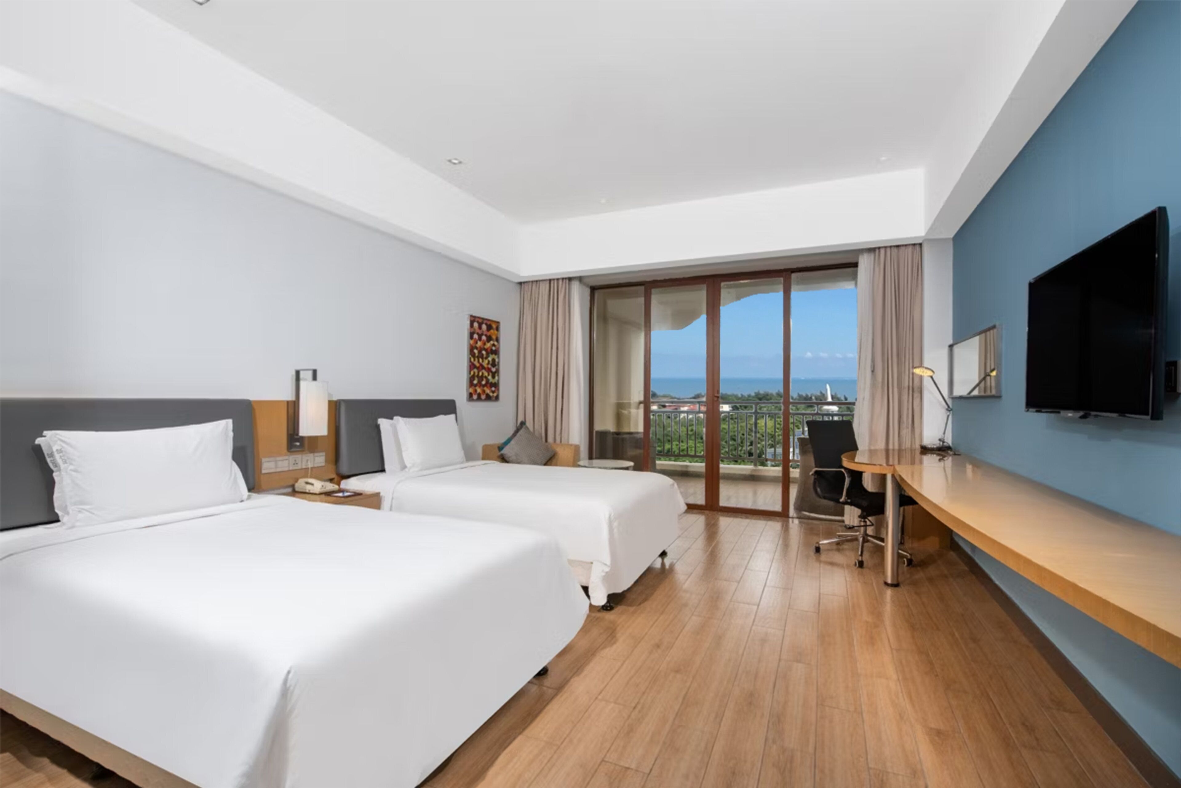 A hotel room with two beds, a desk, a TV, and a balcony with a city view.