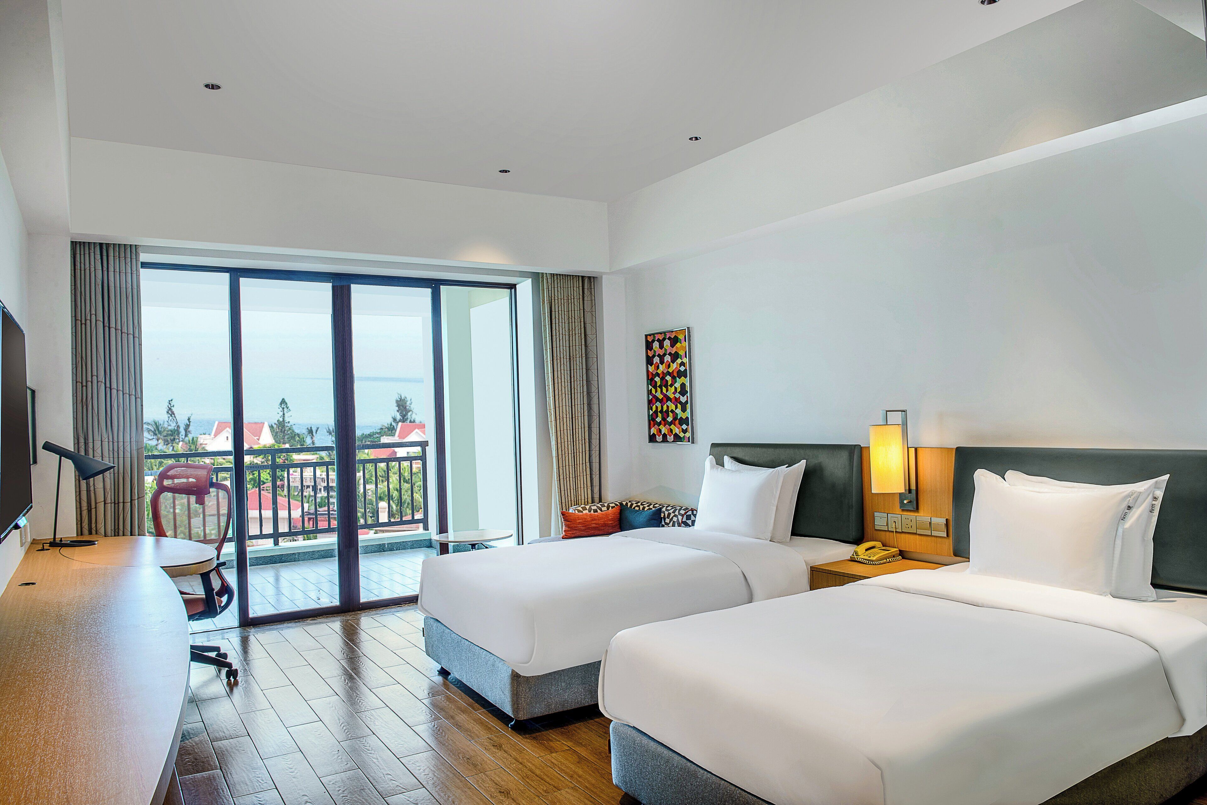 A hotel room with two beds, a desk, a chair, a TV, a balcony with a view, and a painting on the wall.
